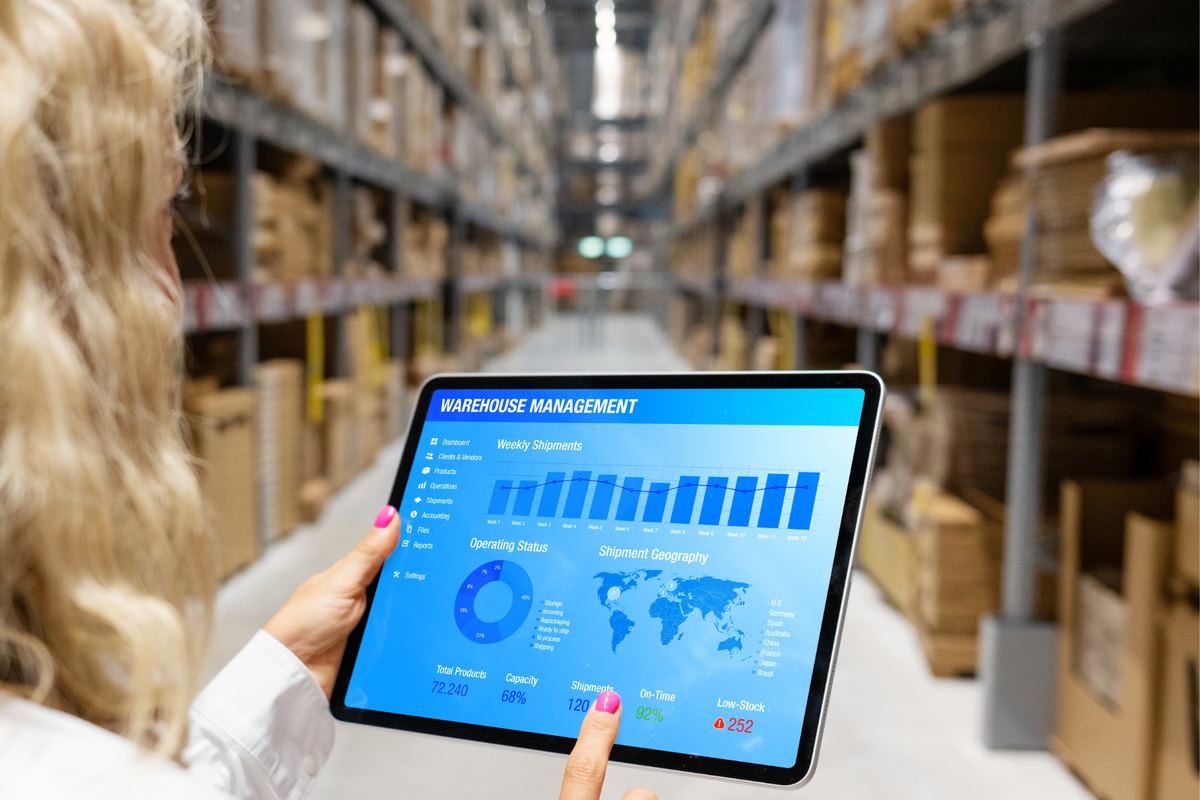 Person holding a tablet displaying a warehouse management dashboard in a warehouse with shelves full of boxes. Blue and white interface.